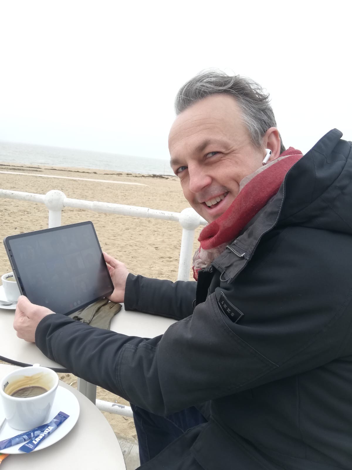 Nicolas Van Caemerbeke, souriant, attablé en terrasse face à une plage, une tablette entre les mains et un café à côté de lui.