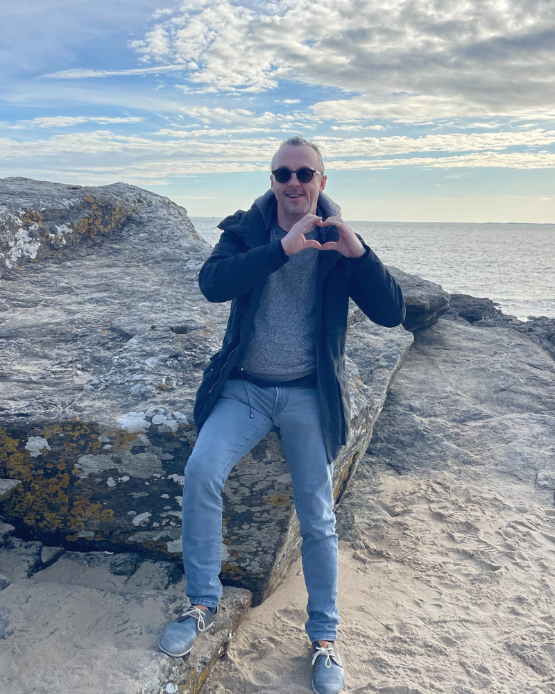 Nicolas Van Caemerbeke, assis sur un rocher face à la mer, forme un cœur avec ses mains.