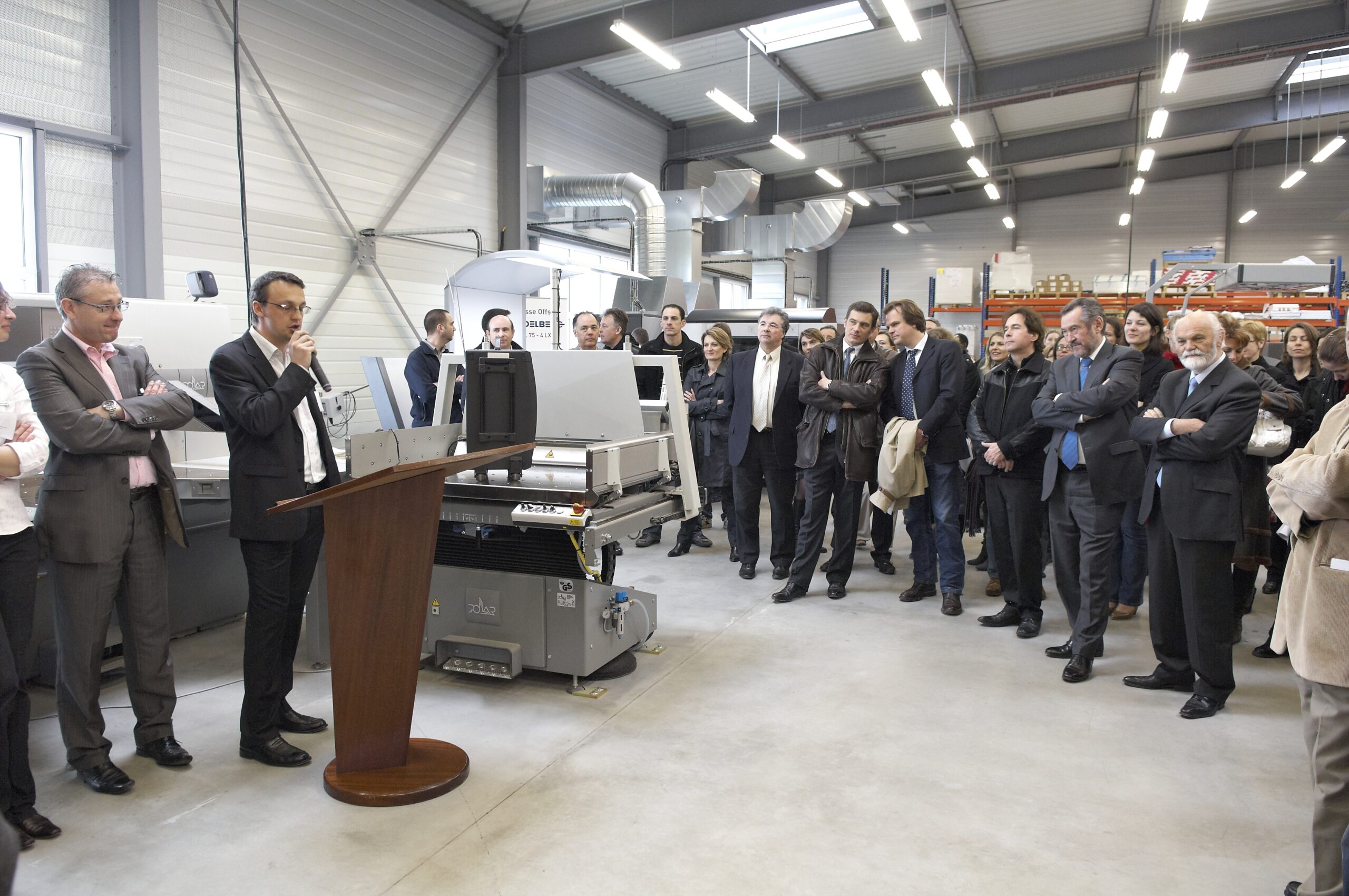 Nicolas Van Caemerbeke, micro en main près d'un pupitre, s'adresse à un groupe réuni dans un atelier d'imprimerie.