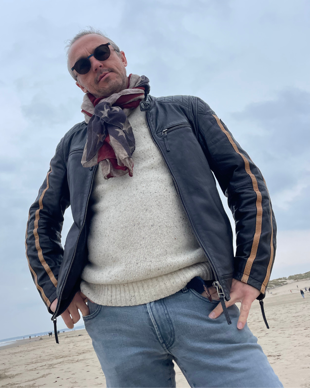 Nicolas Van Caemerbeke, en veste de cuir et lunettes de soleil, les mains dans les poches, photographié en contre-plongée sur une plage.