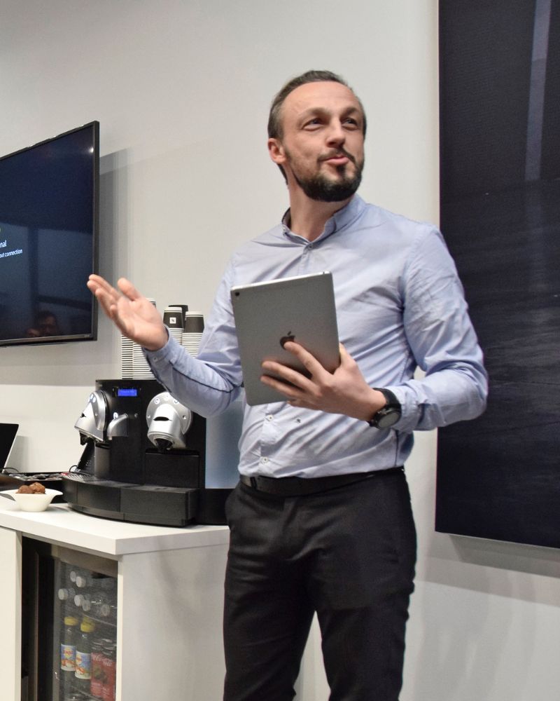 Nicolas Van Caemerbeke, une tablette à la main, s'exprime debout en souriant, devant un écran et une machine à café.
