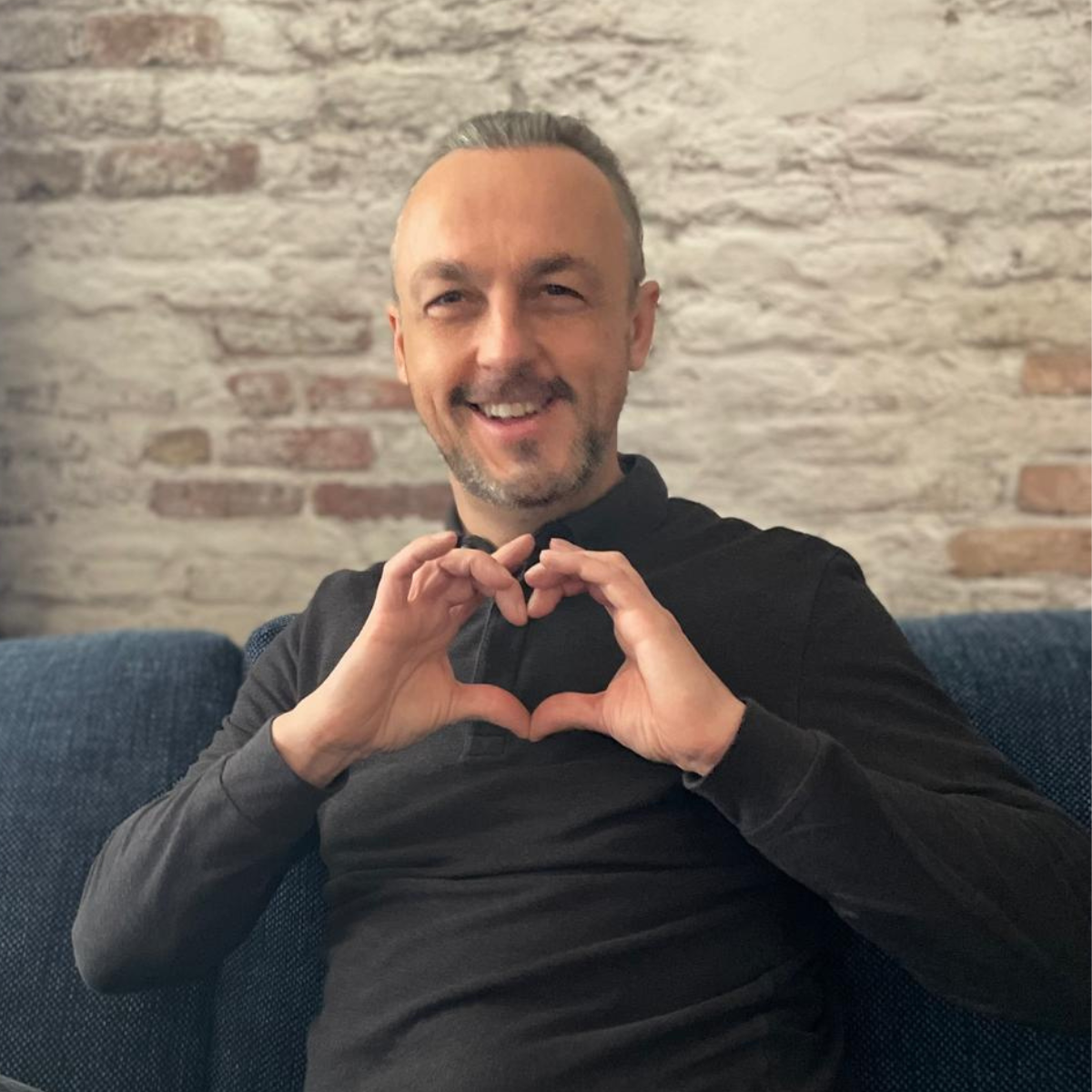 Nicolas Van Caemerbeke, souriant, assis sur un canapé bleu devant un mur de briques blanchies, forme un cœur avec ses mains.