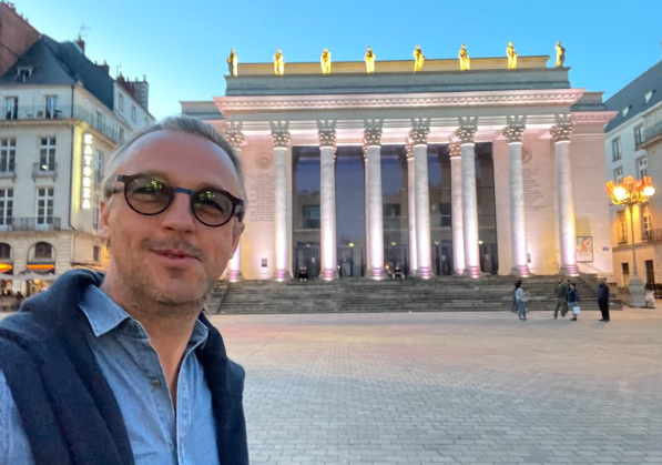 Nicolas Van Caemerbeke, lunettes de soleil, en selfie souriant devant le théâtre Graslin de Nantes illuminé.